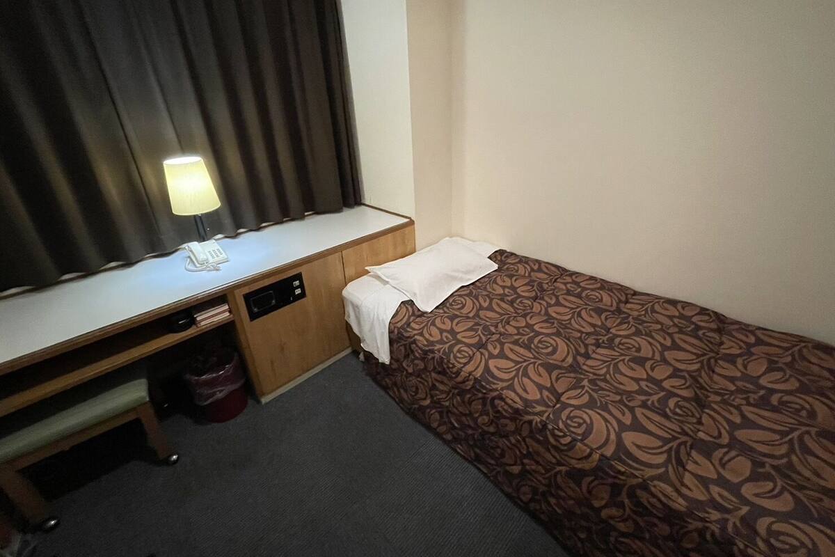 A small hotel room with a single bed, a desk with a lamp, and a window with curtains.