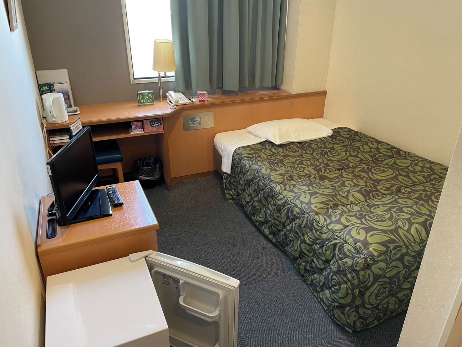 Habitación de hotel pequeña con una cama individual, un escritorio con una computadora portátil, un teléfono y un cesto de basura.