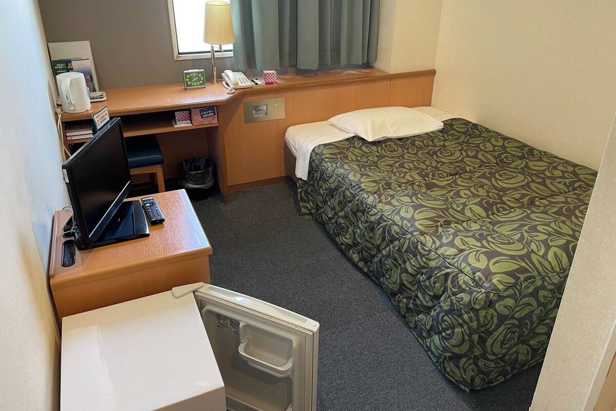 A compact hotel room with a single bed, a desk with a laptop, a telephone, and a small trash bin.
