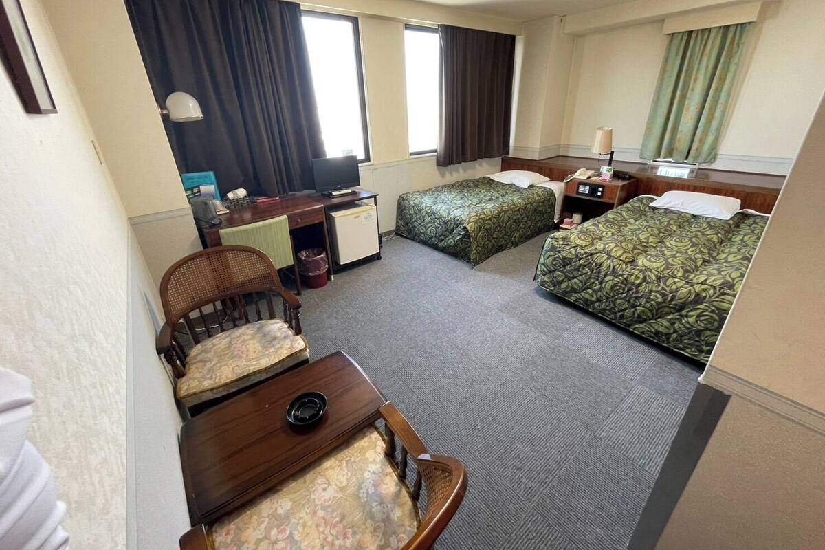 A hotel room with a desk, two beds, a chair, a small table, and a TV.