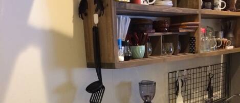 Una cocina con microondas, cafetera y hervidor sobre la encimera. Hay estantes arriba con diversos utensilios de cocina.