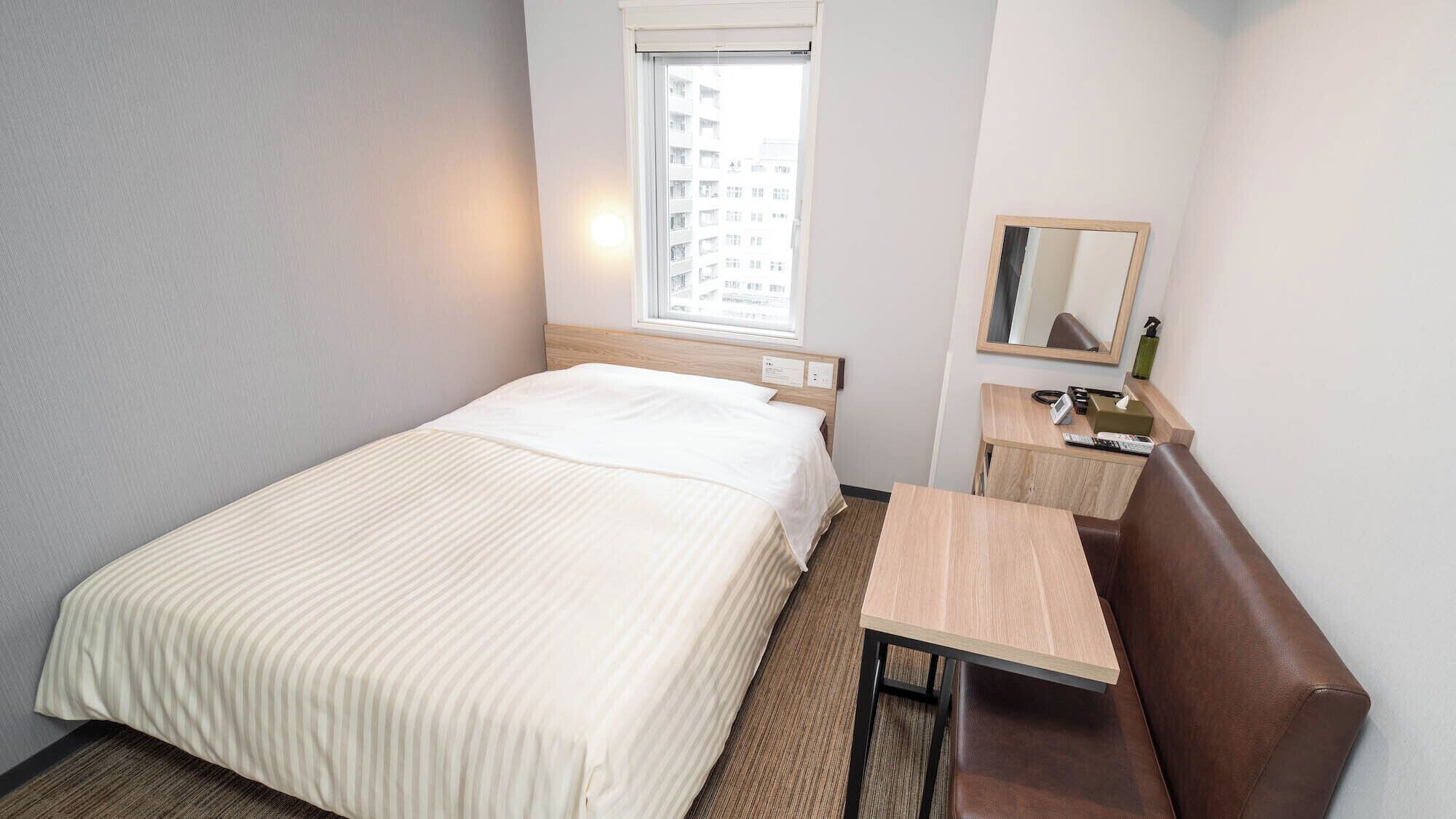 A hotel room with a bed, a small desk, a chair, a mirror, and a window with a view of buildings.