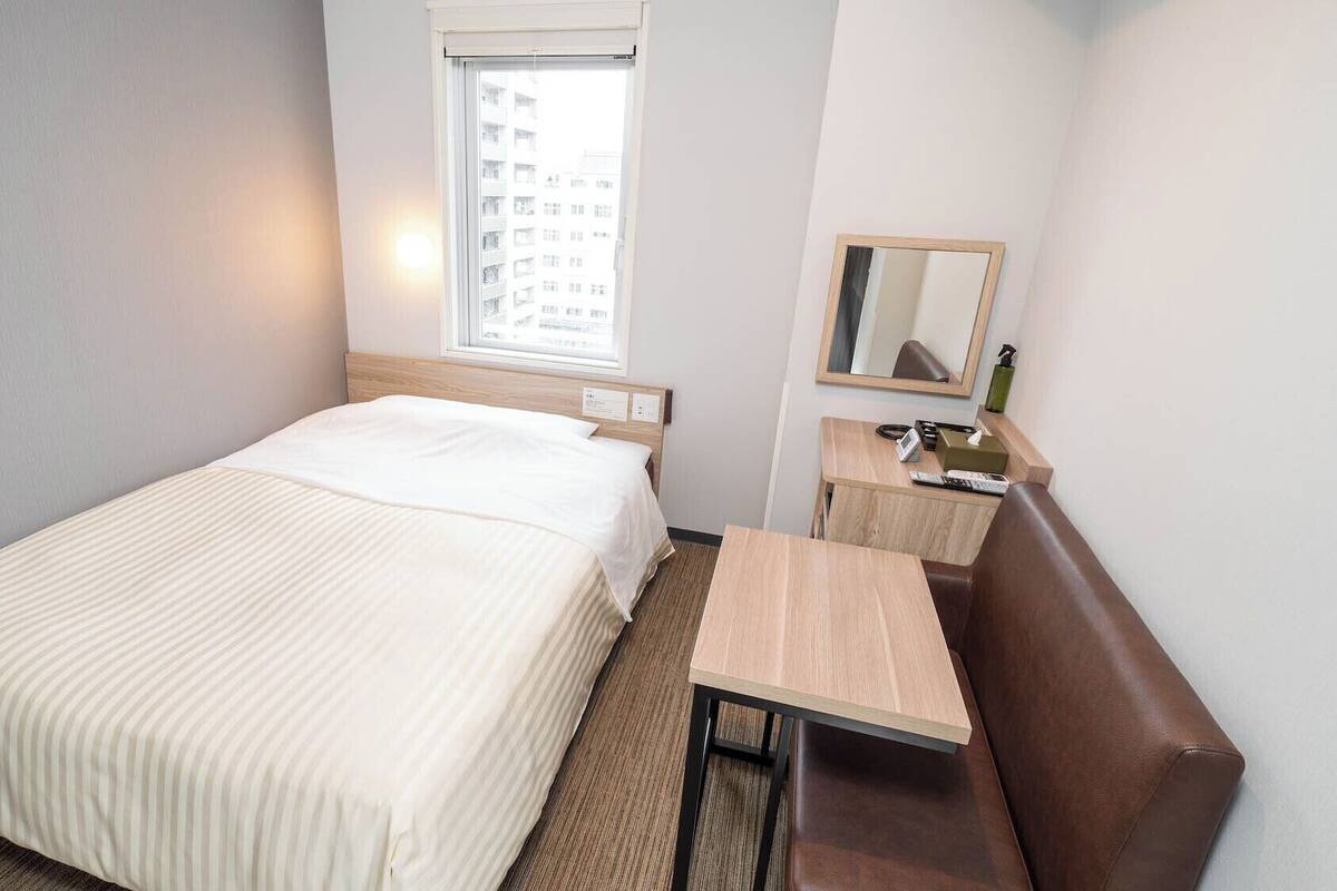 A hotel room with a bed, a small desk, a chair, a mirror, and a window with a view of buildings.