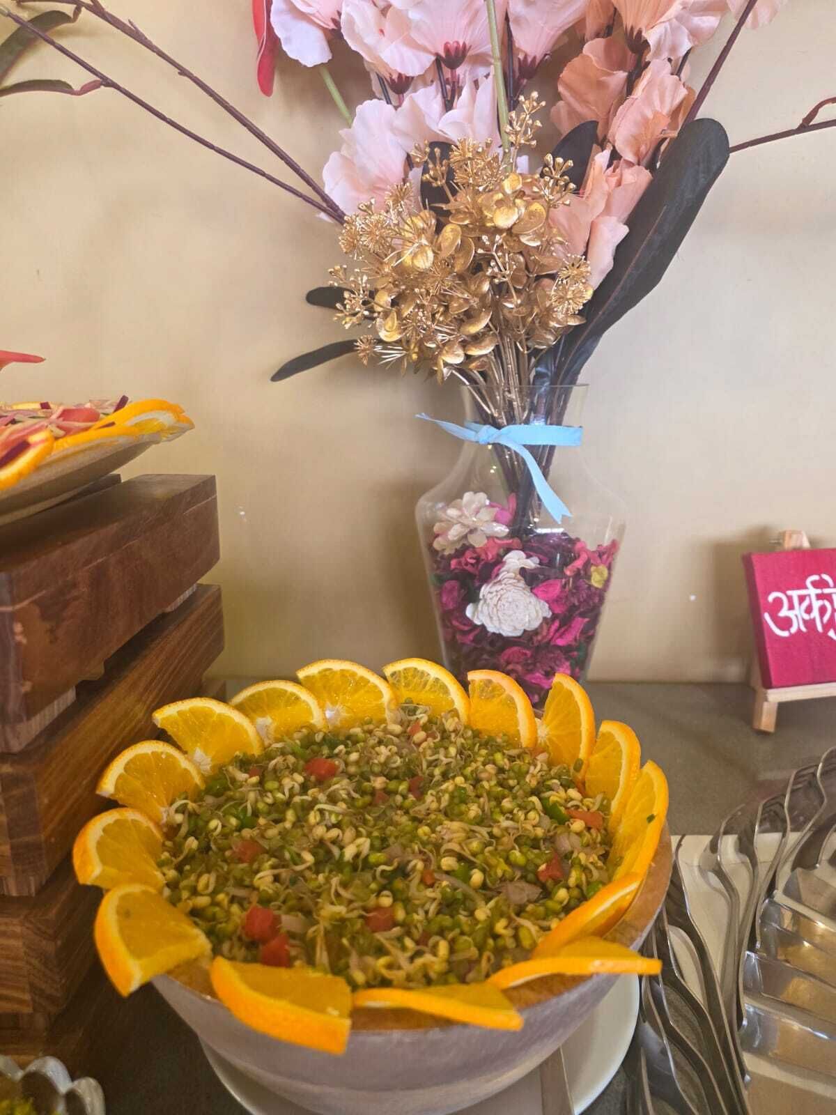 A table with a bowl of salad garnished with orange slices, a vase with pink flowers, and a small sign with text.