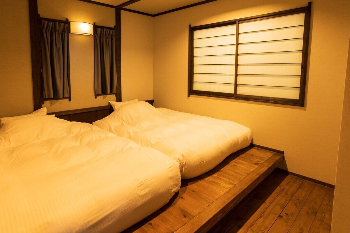A room with two beds, a window with horizontal blinds, a wall-mounted lamp, and a wooden floor.