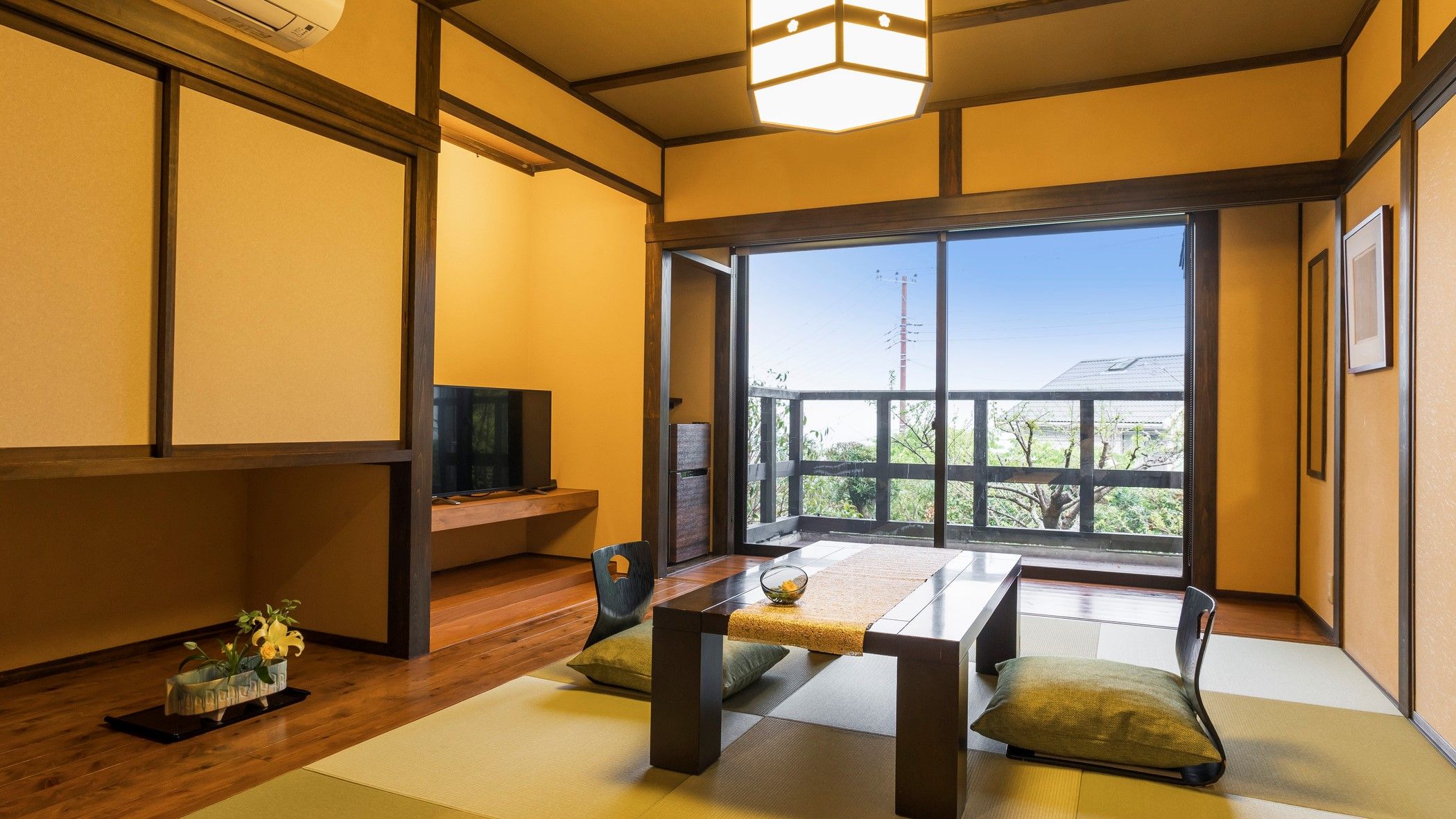 A traditional Japanese-style room with tatami flooring, a low table, cushions, a television, and a large window with a view of greenery.