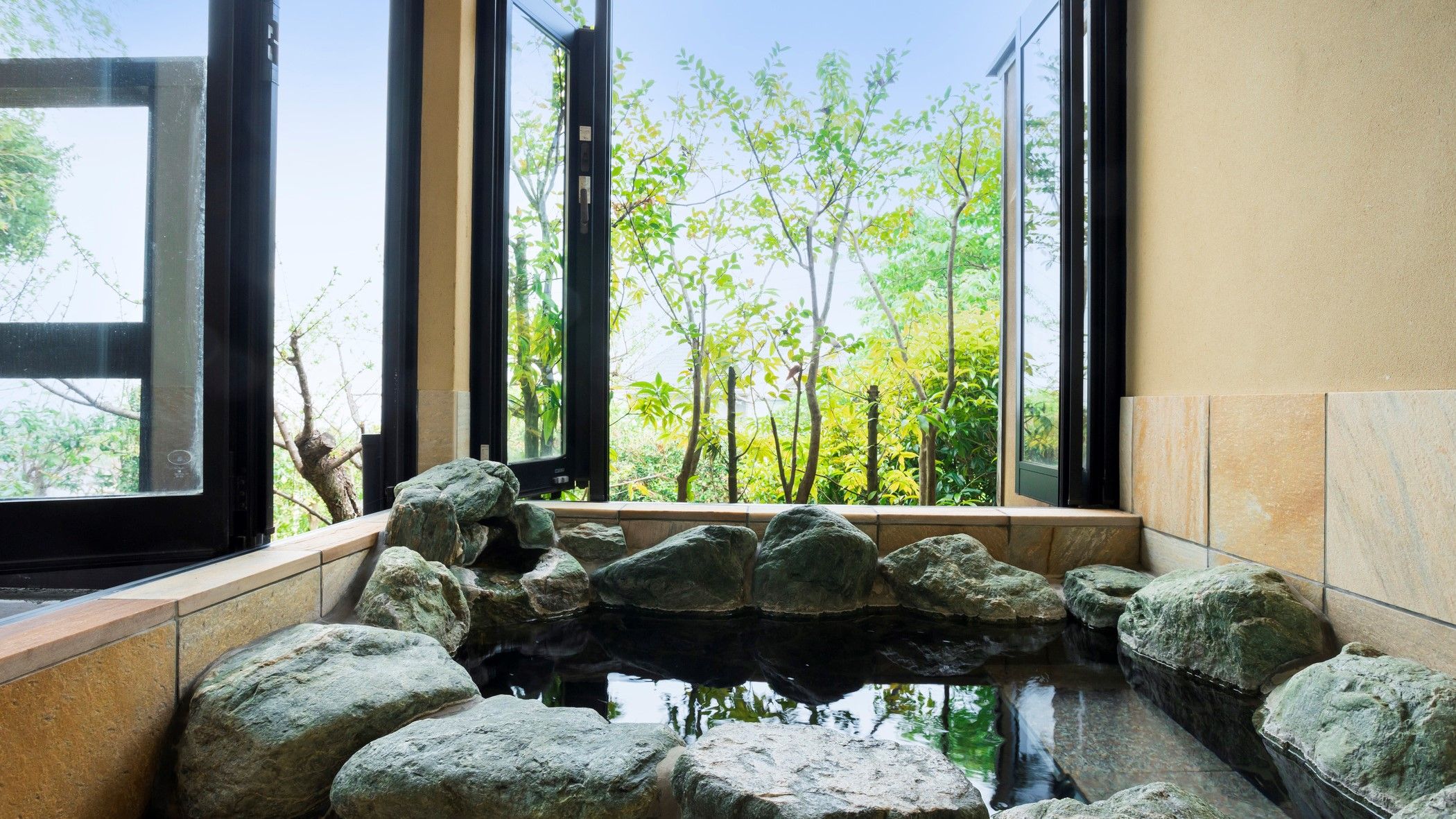 A tranquil indoor water feature with rocks, a view of trees through large windows, and a stone wall.