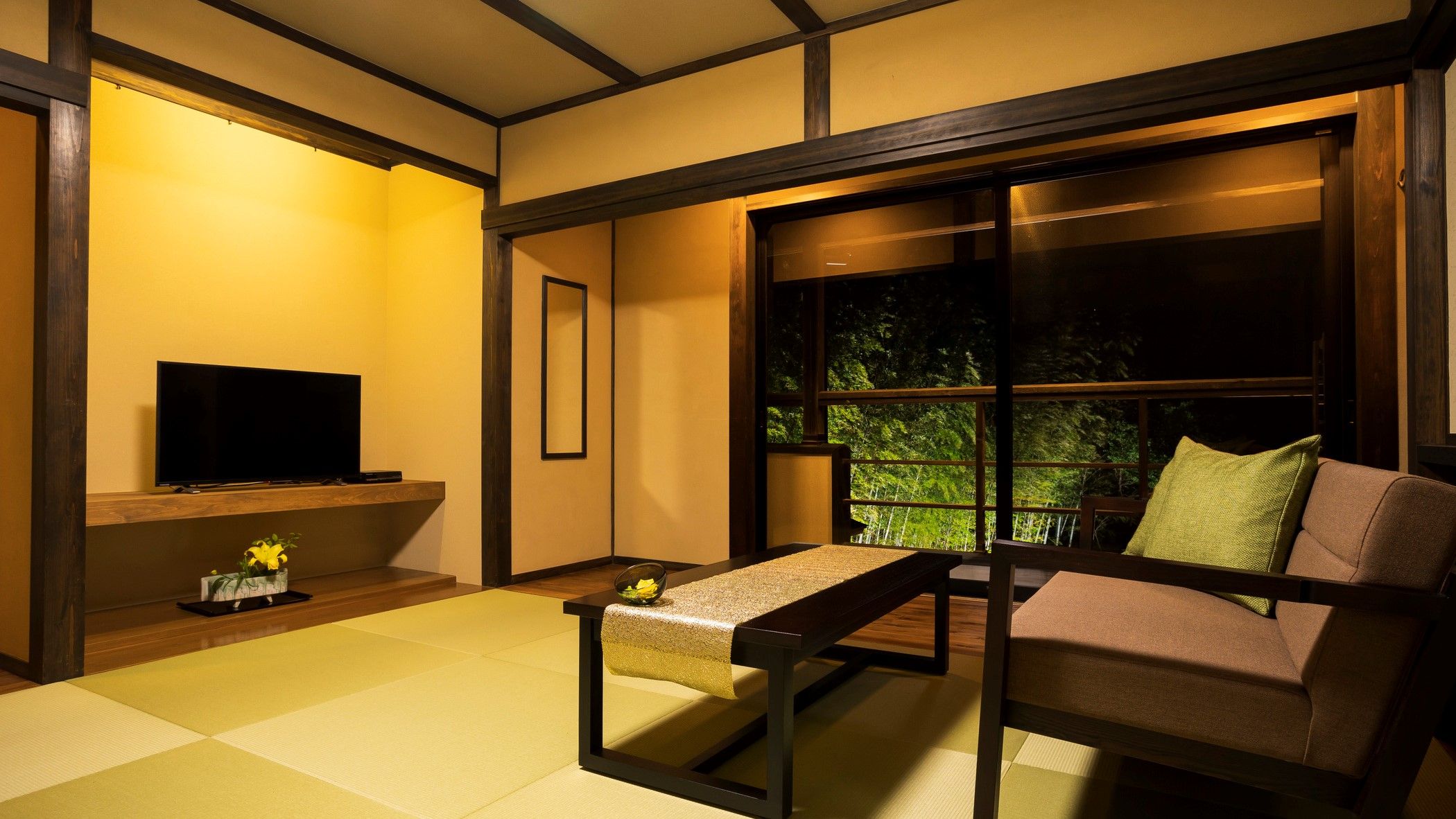 A traditional Japanese-style room with tatami flooring, a low table, a sofa, a TV, and a window with a view of greenery.