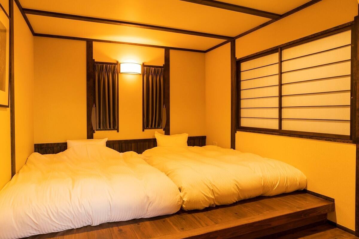 A double bed with a wooden headboard, a bedside table, and a window with horizontal blinds.
