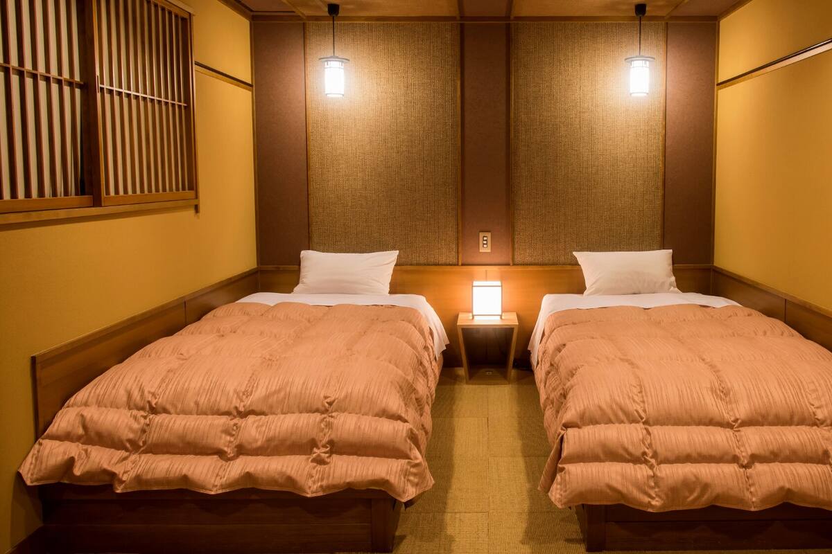 Habitación de hotel con dos camas, una mesita de noche pequeña, una lámpara de pared y una ventana con persianas.