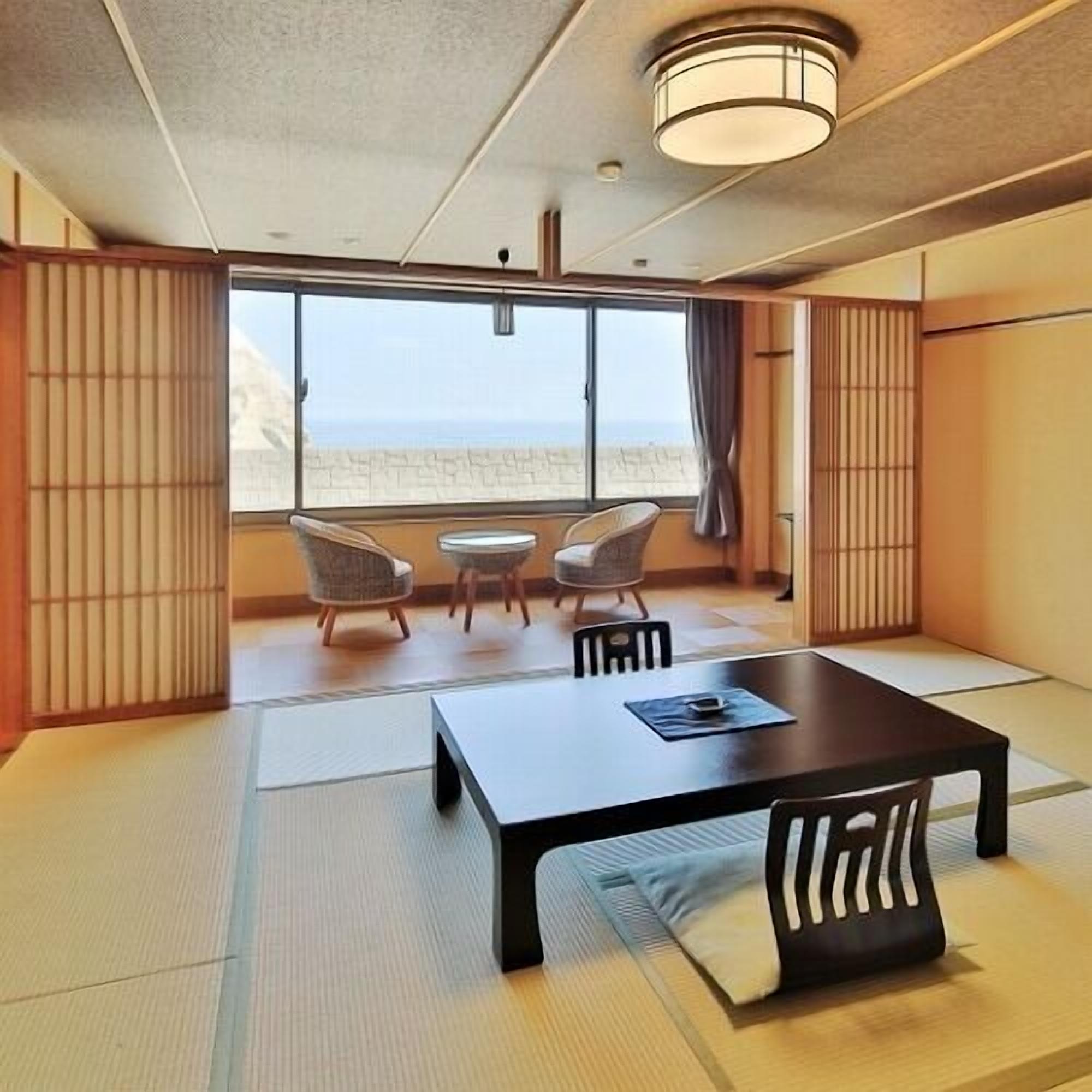 Ein Zimmer mit Tatami-Boden, einem niedrigen Tisch, zwei Stühlen und einem großen Fenster mit Blick auf den Strand.