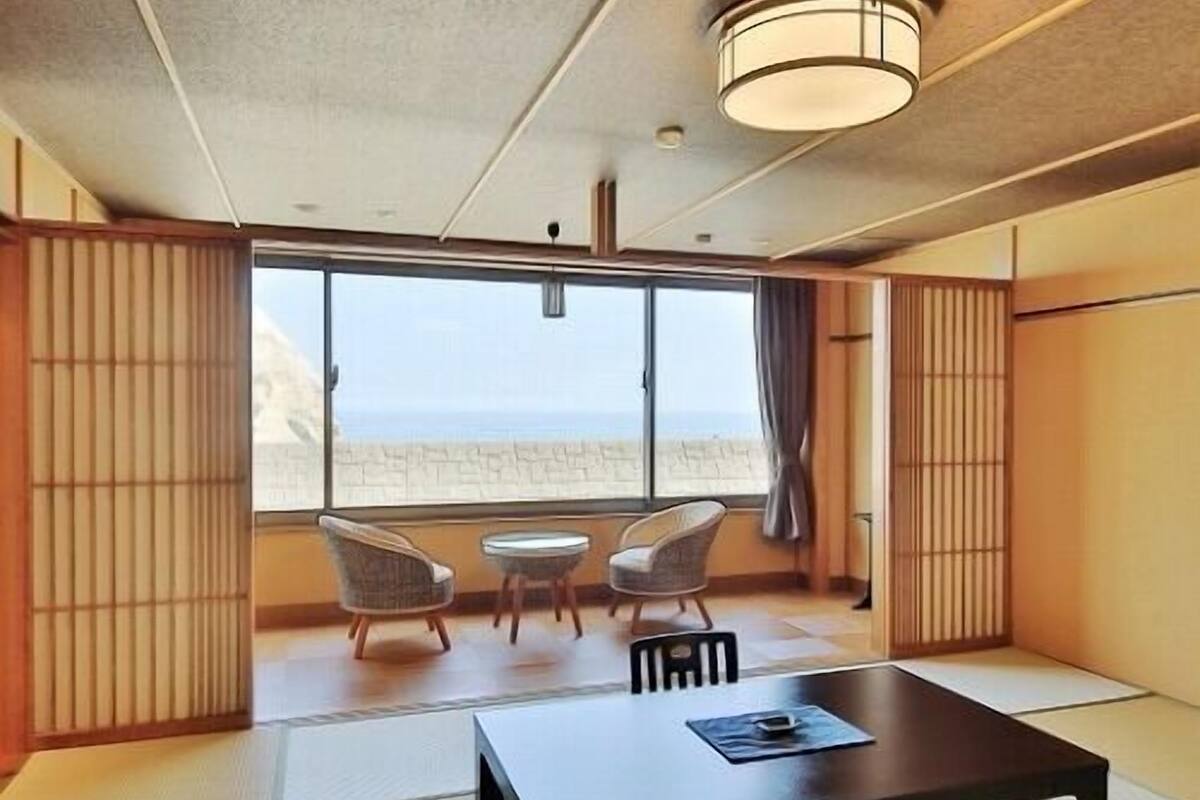 Una habitación con suelo de tatami, una mesa baja, dos sillas y un ventanal con vista a la playa.
