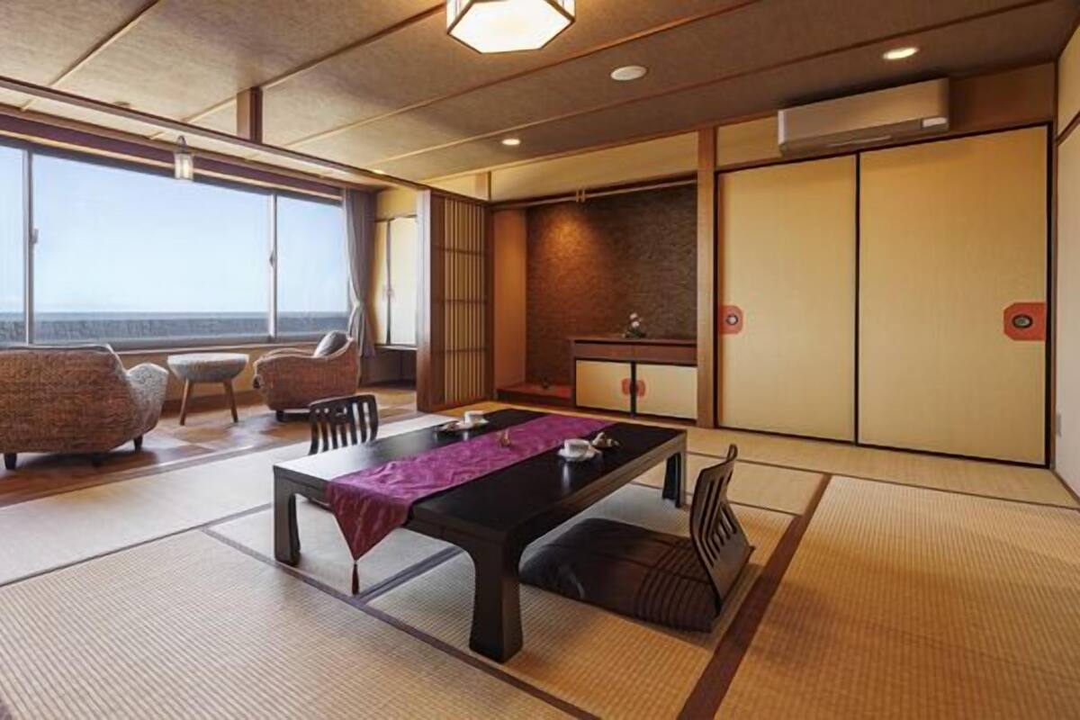Una habitación japonesa tradicional con suelo de tatami, una mesa baja con camino de mesa morado y vistas al mar a través de amplios ventanales.