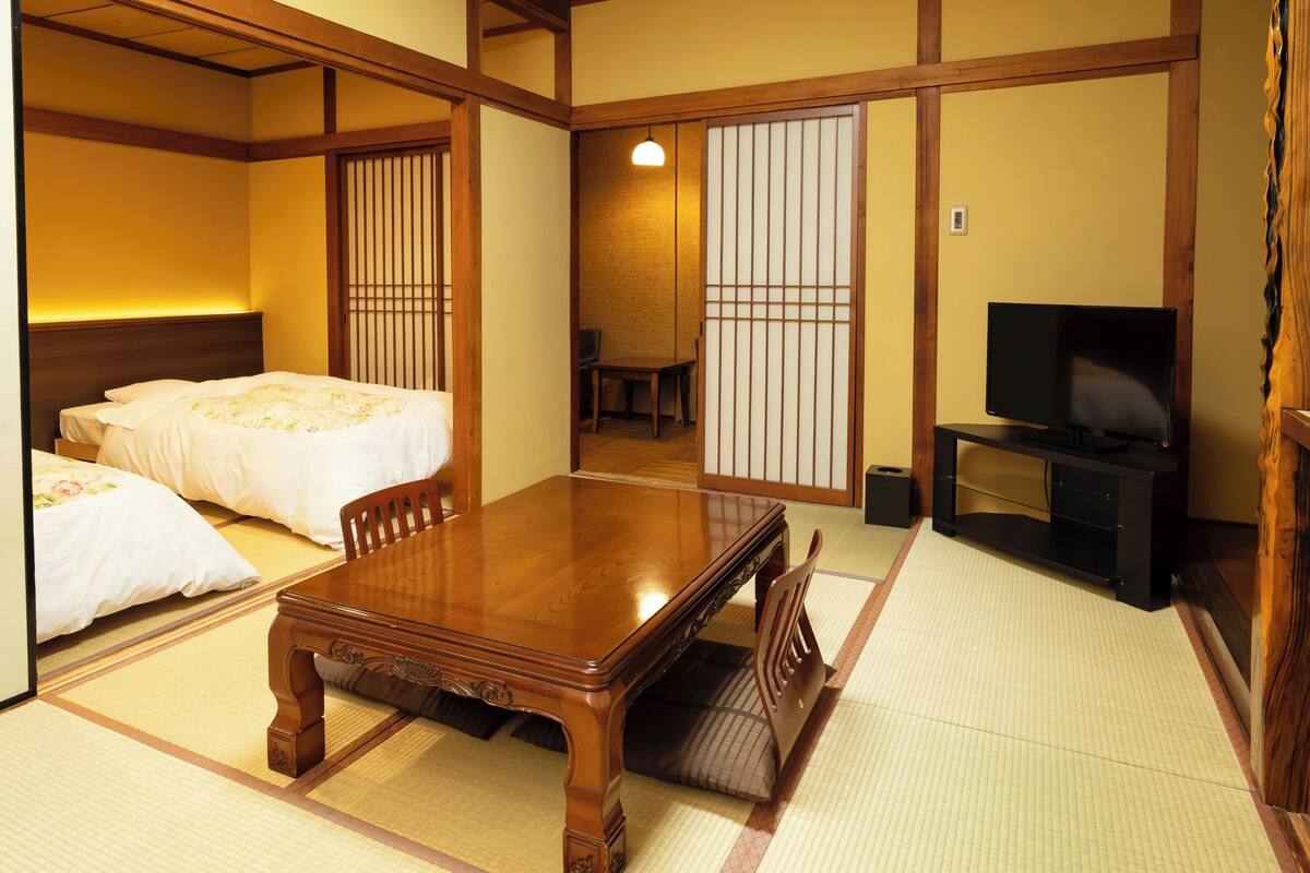 Una habitación japonesa tradicional con suelo de tatami, una mesa de madera, dos camas, un televisor y puertas correderas.
