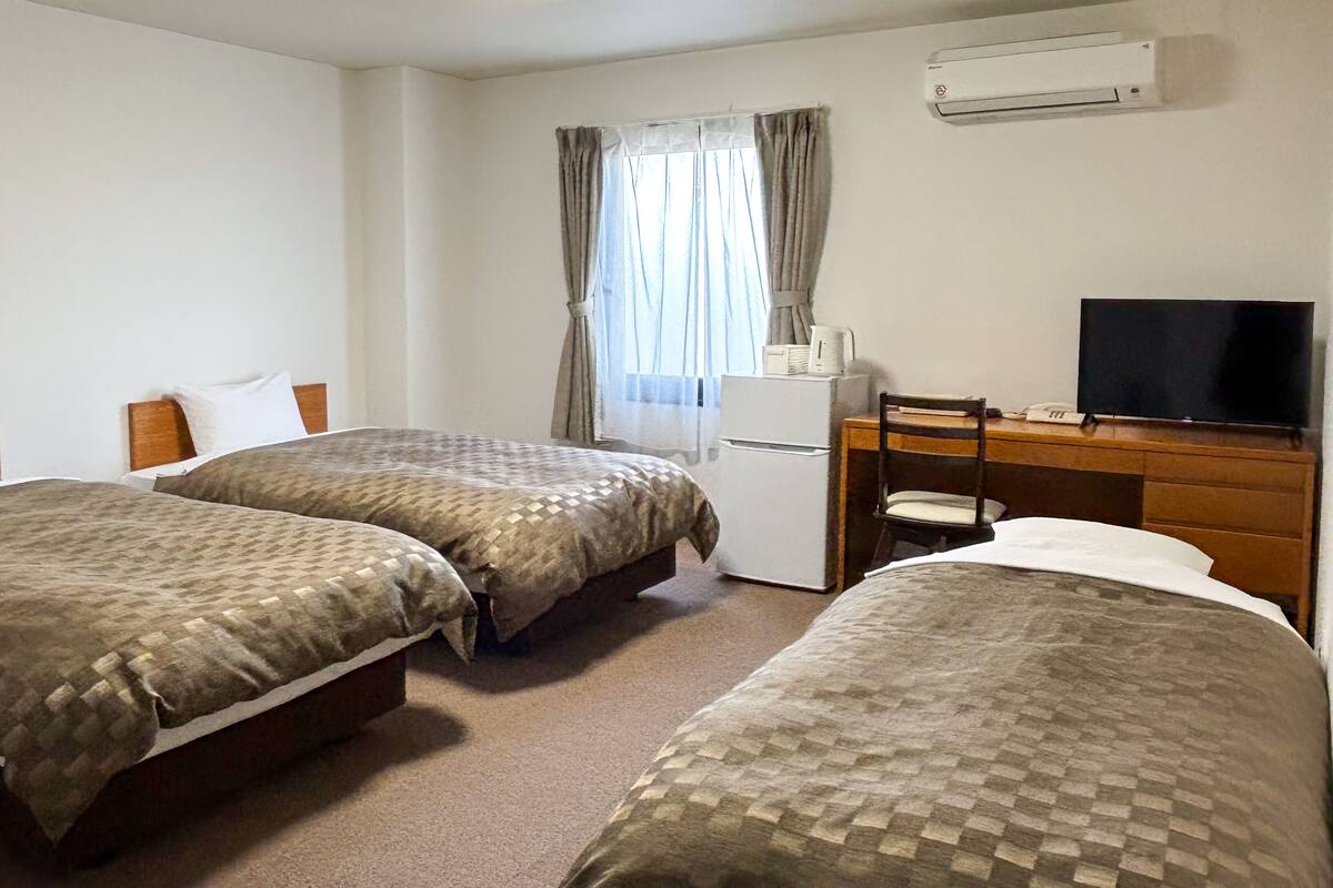 A hotel room with two beds, a desk with a chair, a television, and an air conditioning unit.