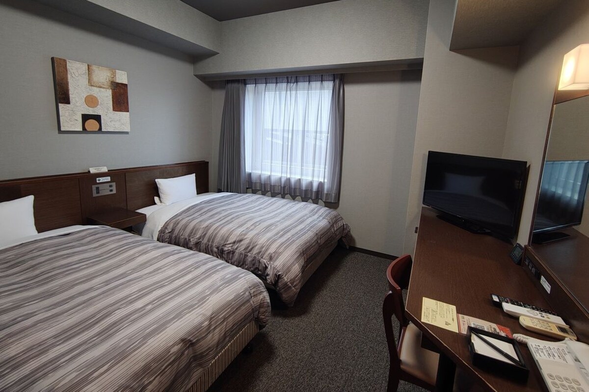 A hotel room with two beds, a desk with a TV, a chair, and a window with curtains.