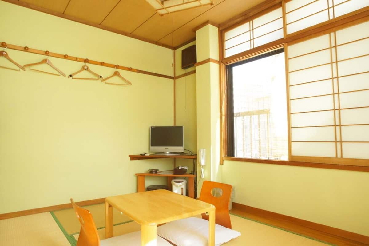 A traditional Japanese room with tatami flooring, a low table with cushions, a television on a shelf, and a sliding shoji screen window.