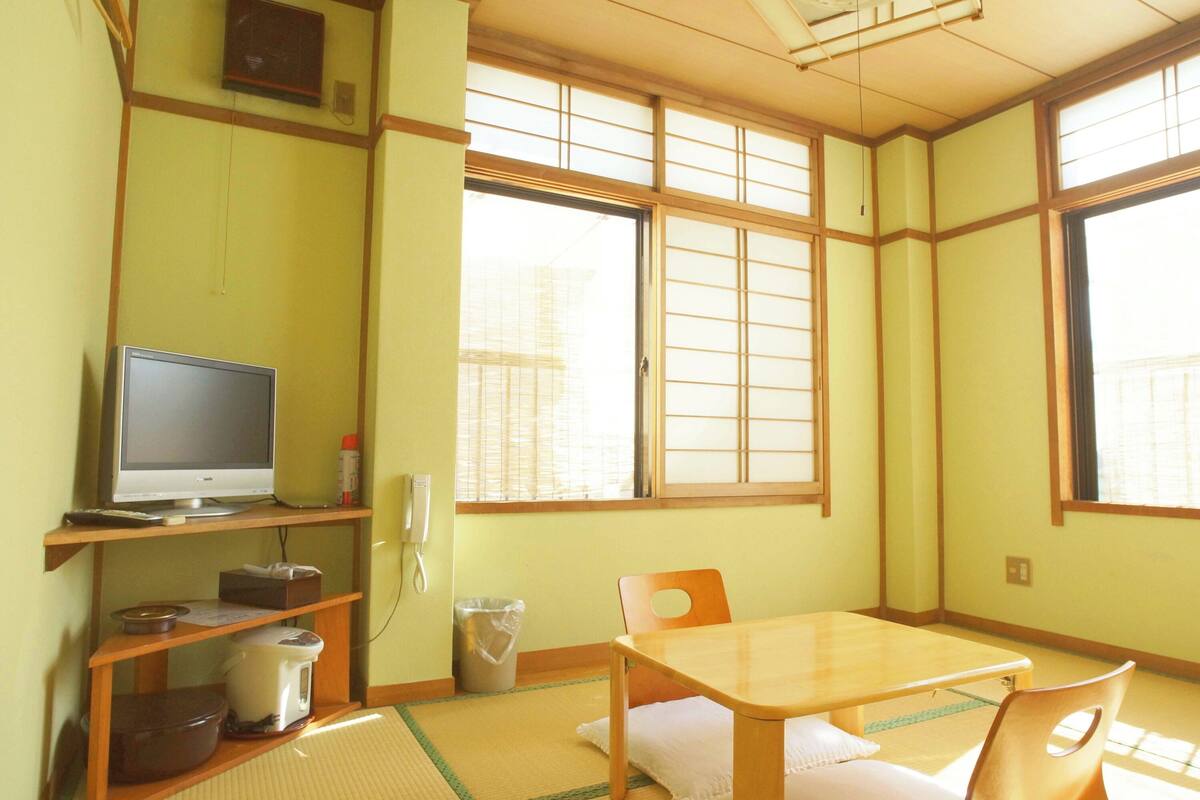 A room with tatami flooring, a television on a shelf, a table with chairs, and large windows with wooden frames.
