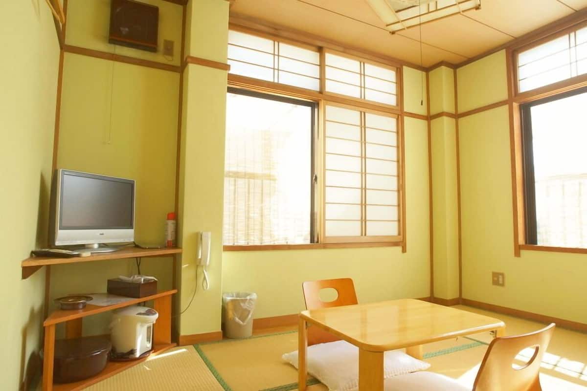 A traditional Japanese room with tatami flooring, a television on a shelf, a table with chairs, and large windows.