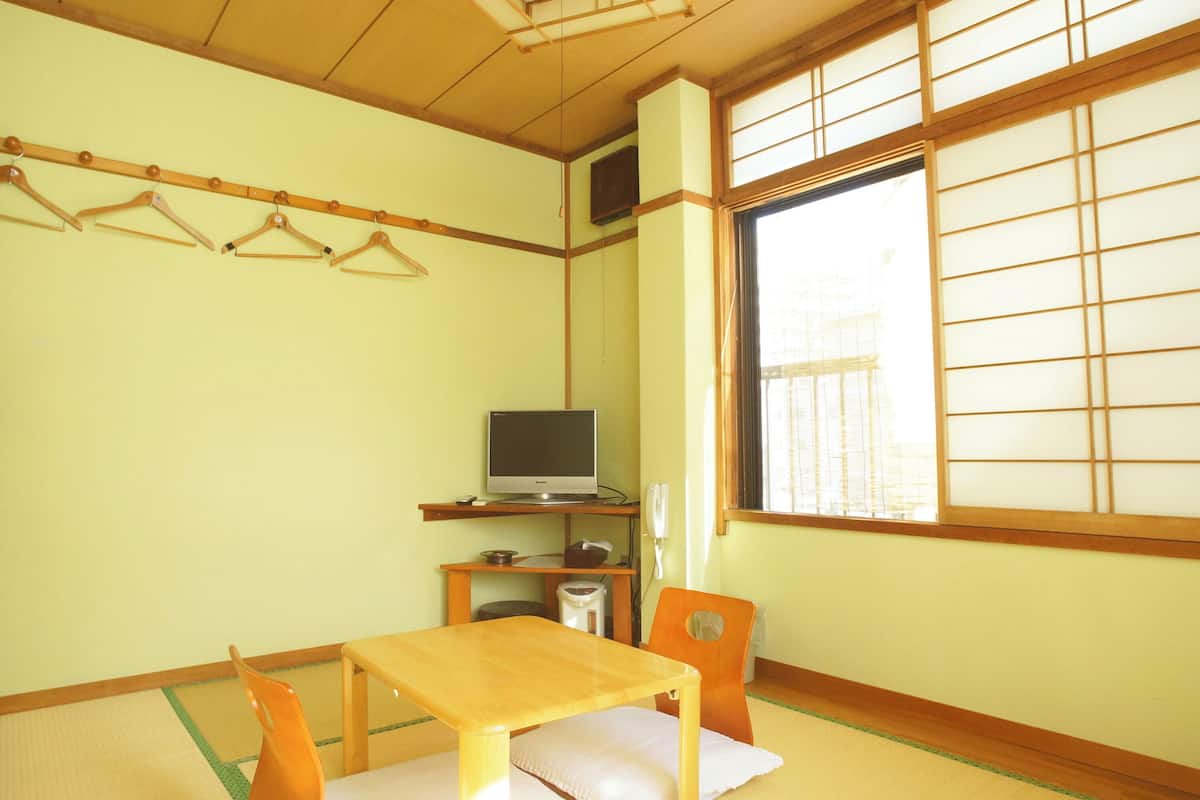 A traditional Japanese room with tatami flooring, a low table with a cushion, a television on a shelf, and a sliding shoji screen.