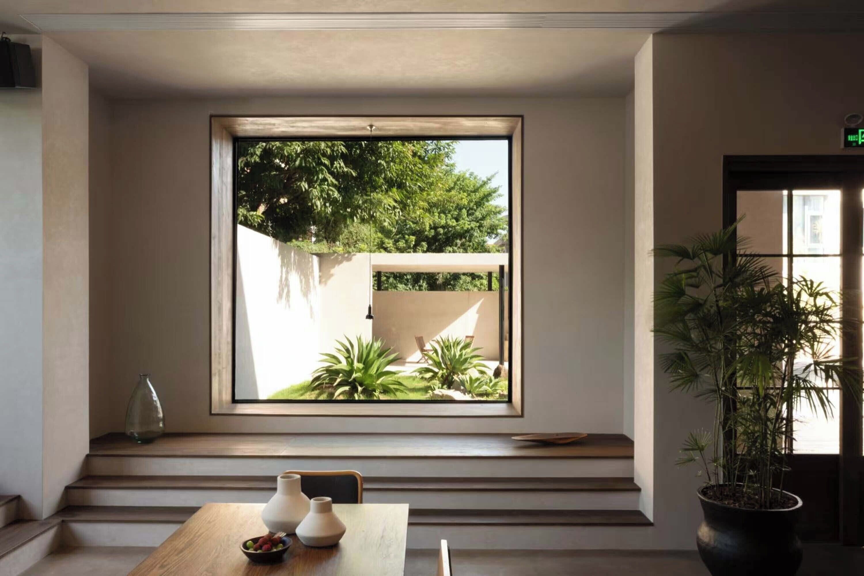 Una habitación con un ventanal grande con vista a un jardín, una mesa de madera con un jarrón y un cuenco, y una planta en maceta a la derecha.