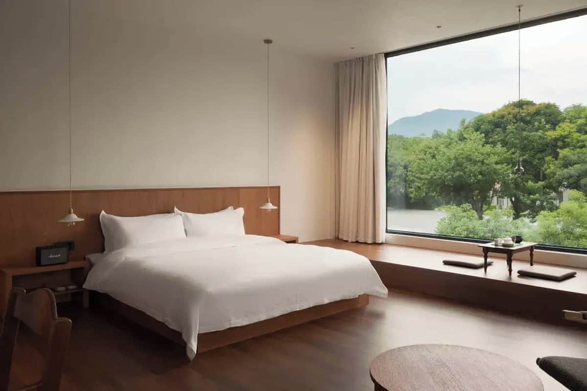 Ett modernt hotellrum med en stor säng, ett sängbord, en stol och utsikt  över ett frodigt, grönskande landskap genom ett stort fönster.