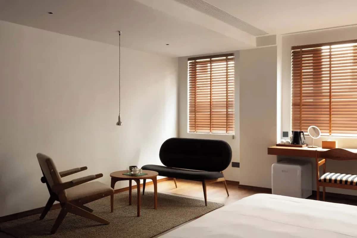 Ett modernt hotellrum med en säng, ett skrivbord med en dator, en stol, ett litet bord och ett fönster med persienner.