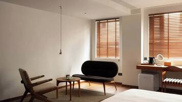 A modern hotel room with a bed, a desk with a computer, a chair, a small table, and a window with blinds.