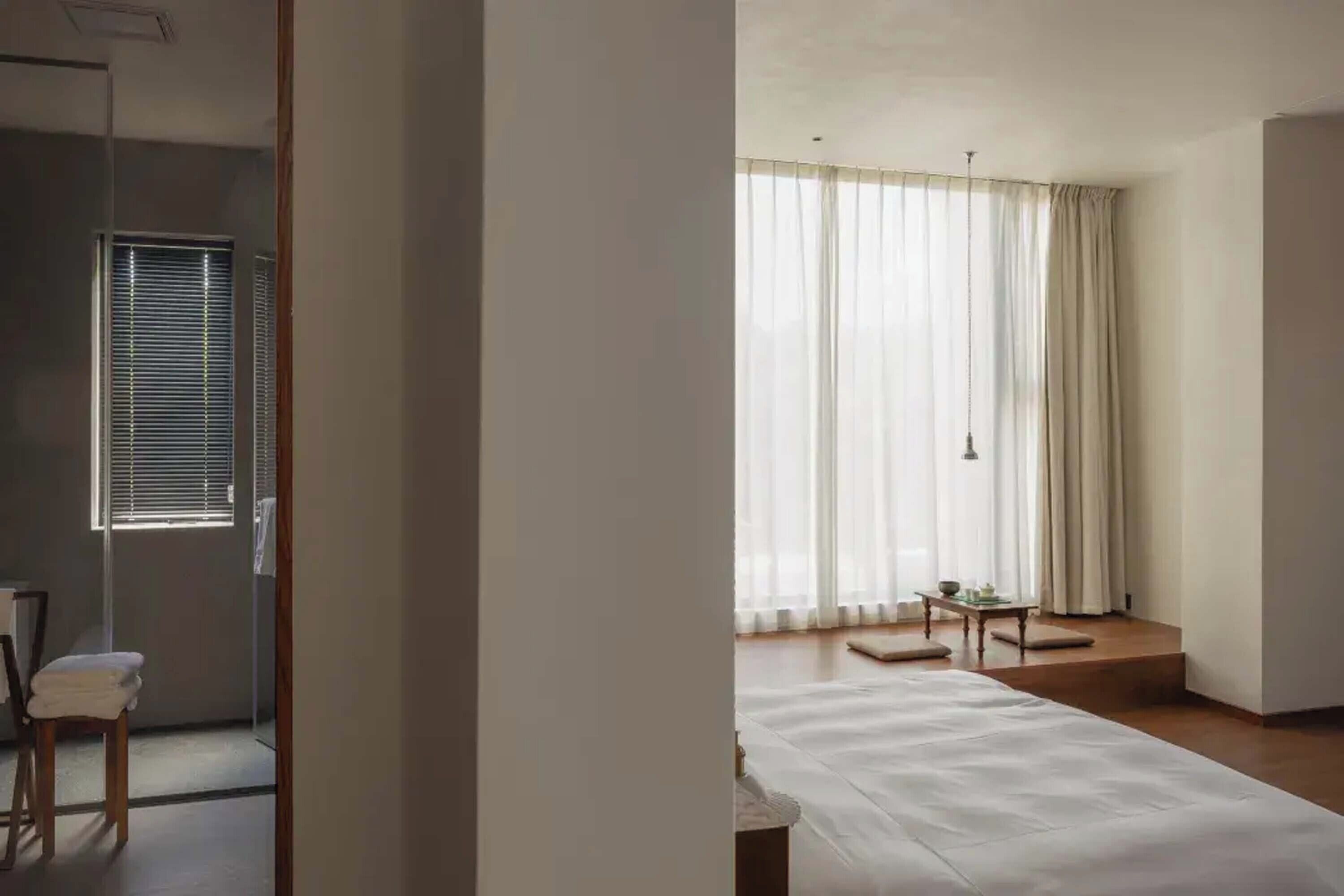 Quarto de hotel moderno com uma cama grande, uma mesinha e uma janela com cortinas.