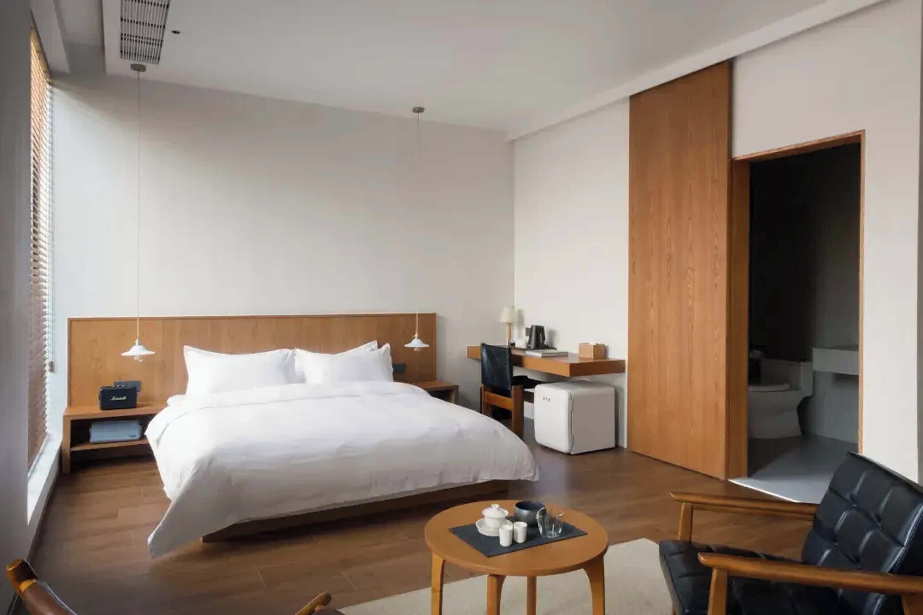 Una habitación de hotel moderna con una cama grande, un escritorio, una silla, una mesita y un baño visible a través de una puerta abierta.