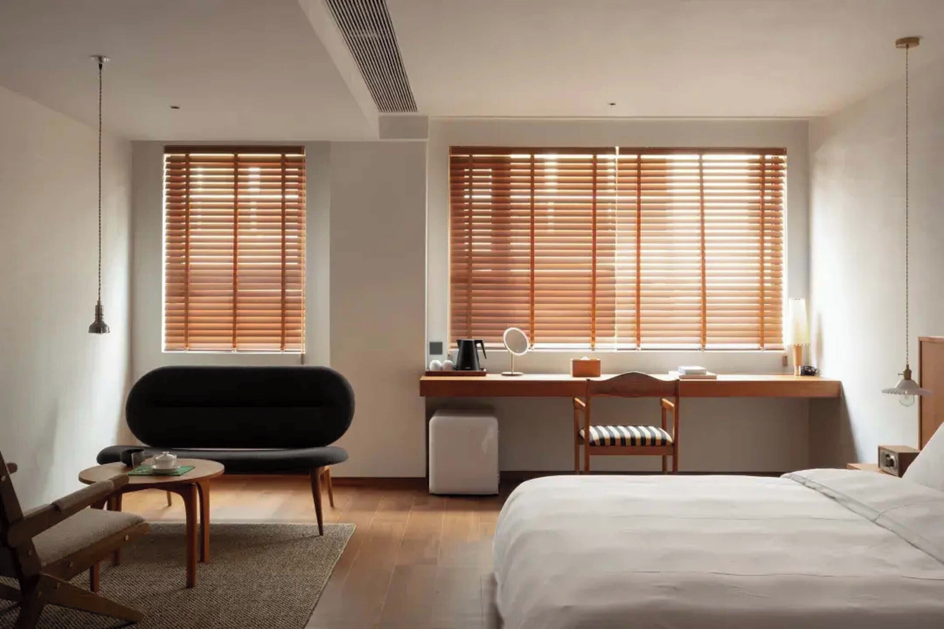 Une chambre d’hôtel moderne comprenant un lit, un bureau avec une chaise, une petite table et deux fenêtres équipées de stores.