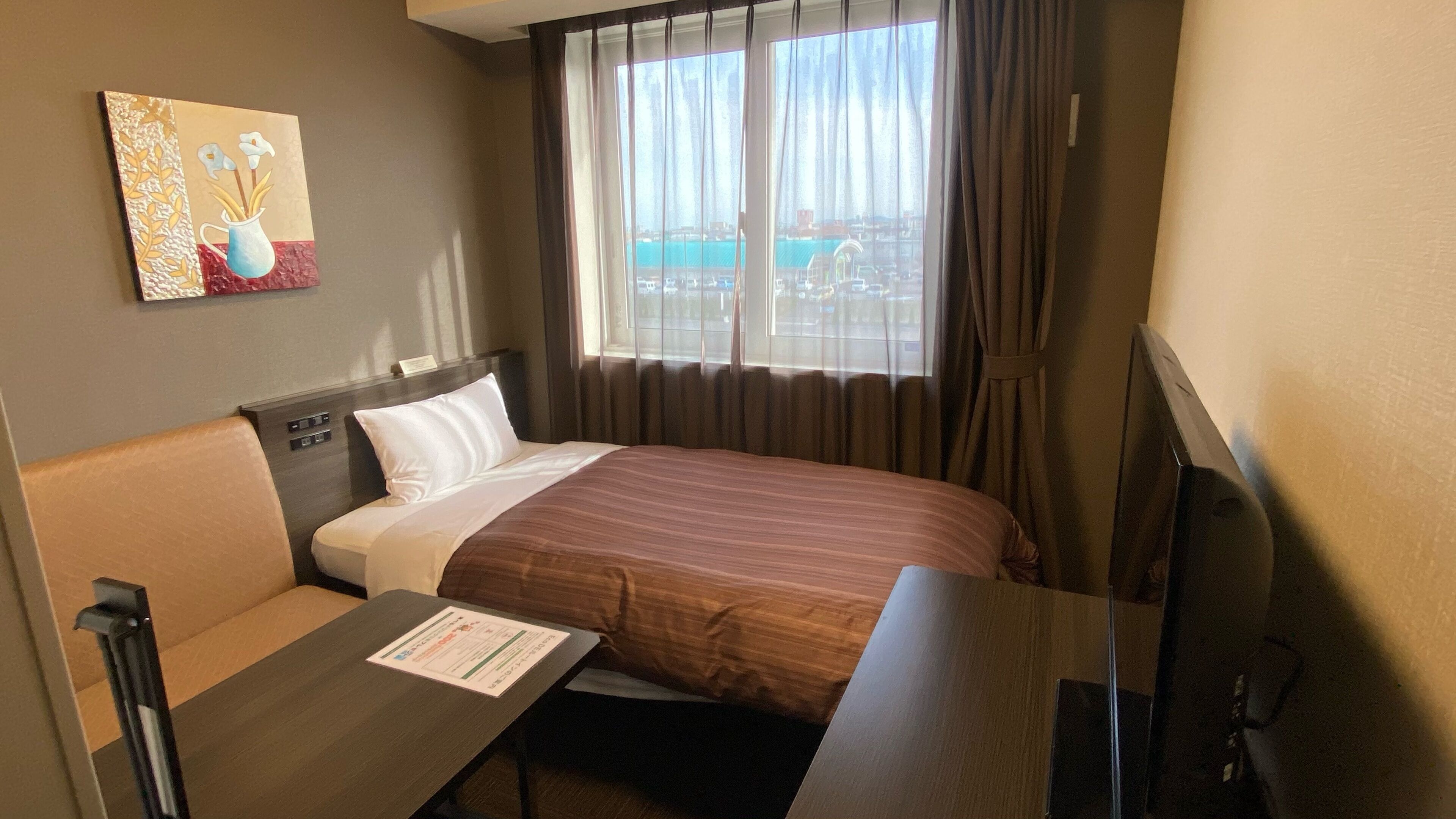 A hotel room with a bed, a desk, a chair, a TV, a painting on the wall, and a window with a view.