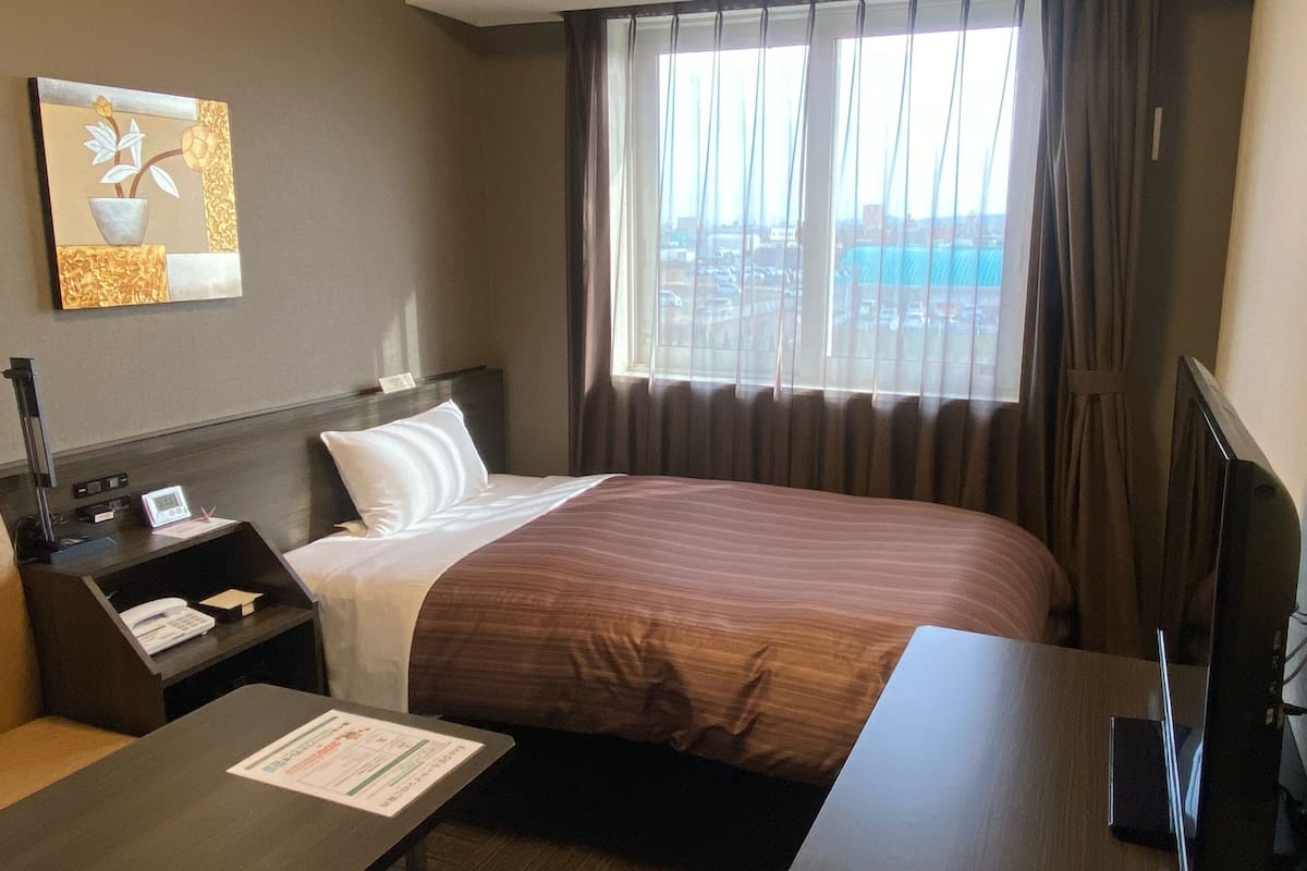 A hotel room with a bed, a desk, a chair, a TV, and a window with curtains.