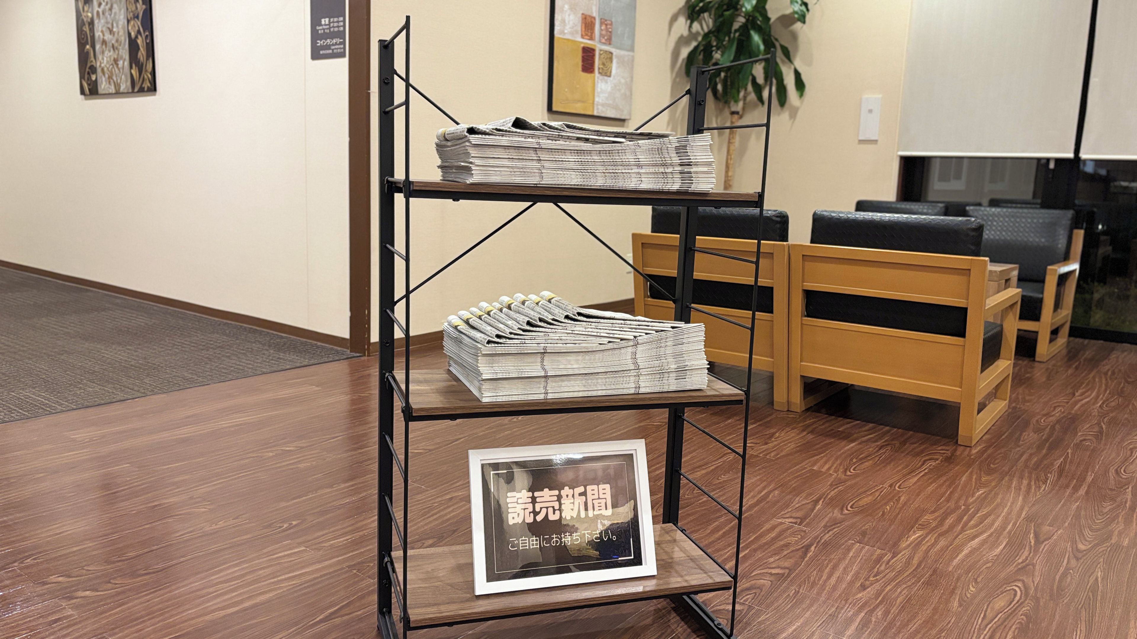 A reception desk with a sign, a stack of newspapers, and a plant in the background.