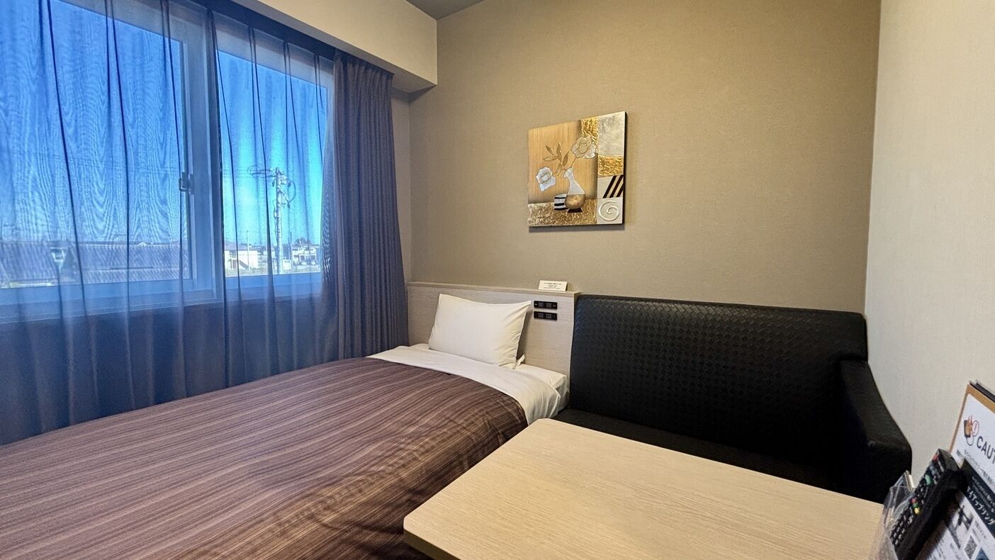 Quarto de hotel com cama, sofá, janela com cortinas e uma obra de arte na parede.