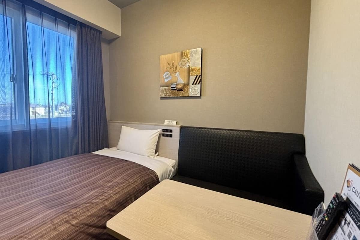 A hotel room with a bed, a sofa, a window with curtains, and a wall-mounted artwork.
