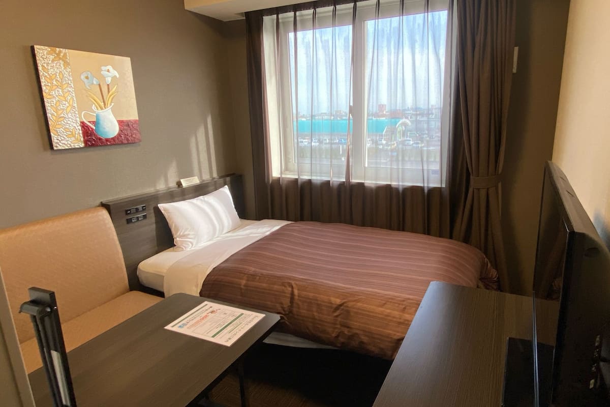 A hotel room with a bed, a desk, a chair, a TV, a painting on the wall, and a window with a view.