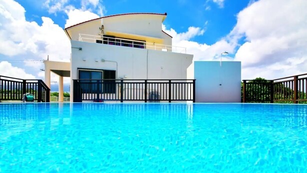 A swimming pool with a white building in the background. - Villa Luxe (Nakijin)