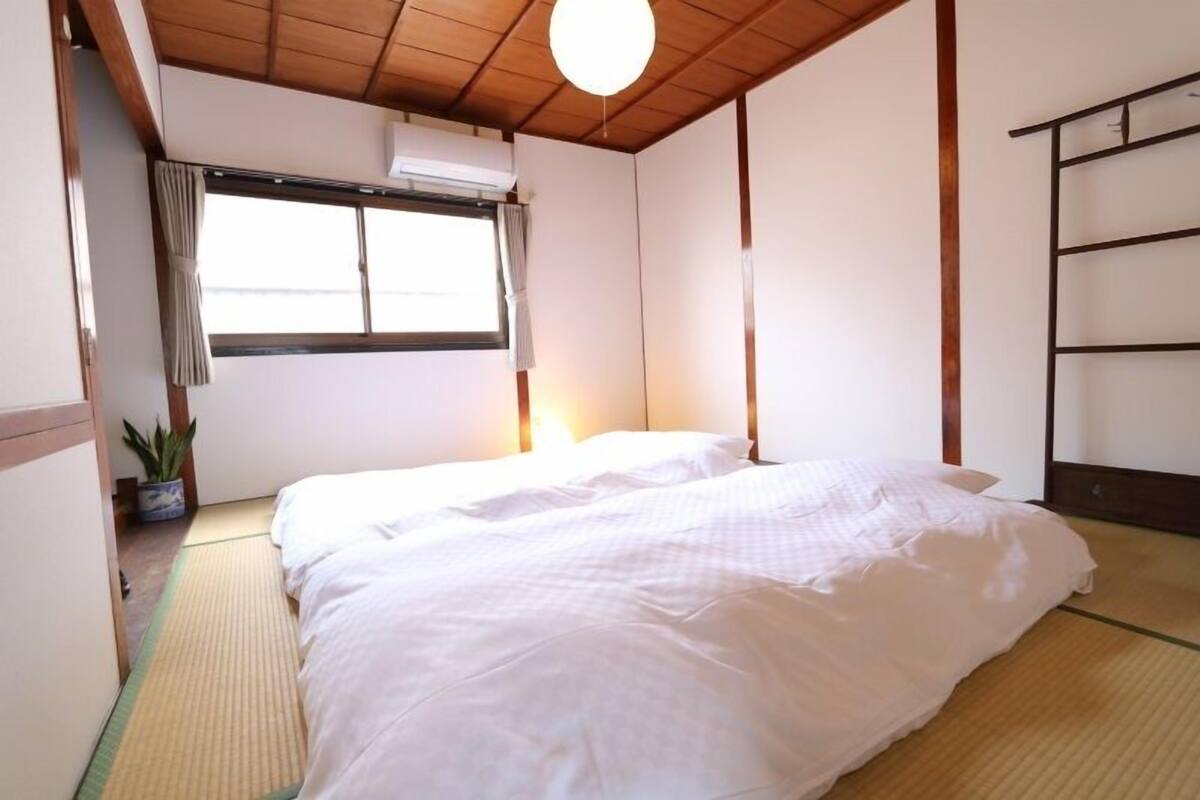 A room with tatami flooring, a bed with white bedding, a window with curtains, an air conditioning unit, and a shelf with items.