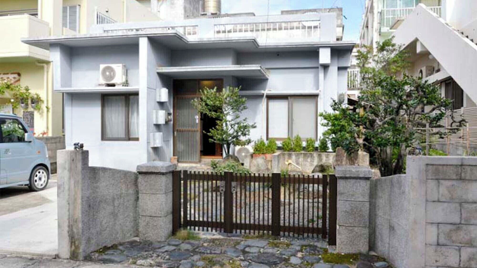 Un moderno edificio residenziale su due piani con tetto piatto, ingresso con cancello e un piccolo giardino con piante in vaso.