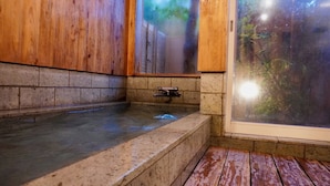 A stone bathtub with a wooden bench, a metal faucet, and a glass door leading to a garden. - Pension Twilight (Nasu)
