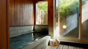 A wooden interior with a stone-lined hot tub, a window with blinds, and a view of greenery outside. - Pension Twilight (Nasu)
