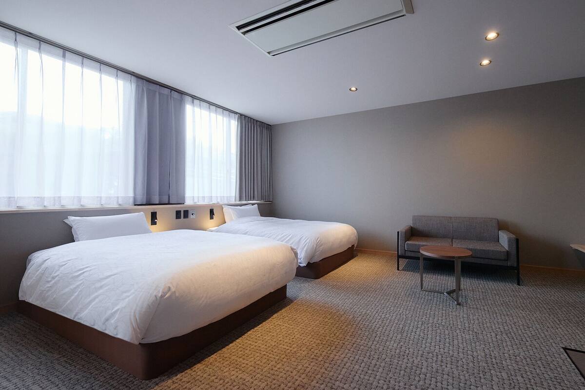 A hotel room with two beds, a sofa, a small table, a TV, and an air conditioning unit.