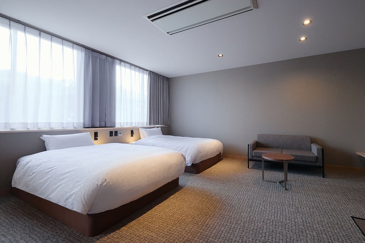 A hotel room with two beds, a sofa, a small table, a TV, and an air conditioning unit.