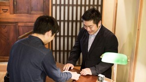 Two individuals seated at a wooden table in a room with traditional wooden paneling and a green desk lamp. - Araiya (Tokyo)
