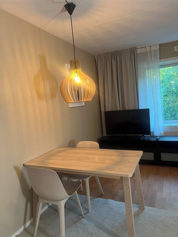 Una sala de estar moderna con una mesa de comedor de madera, sillas blancas, un televisor sobre un soporte negro y una lámpara colgante original.
