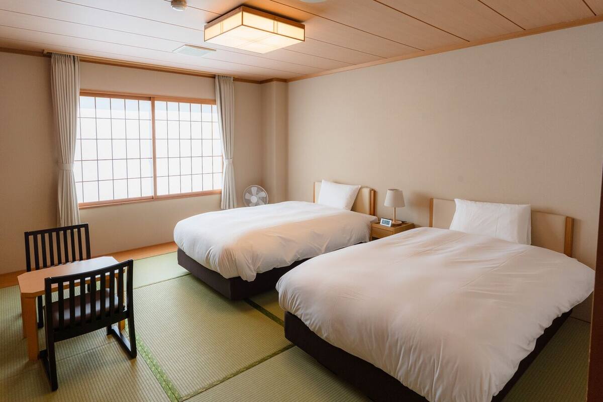 A hotel room with two beds, a desk, a chair, a fan, and a window with curtains.
