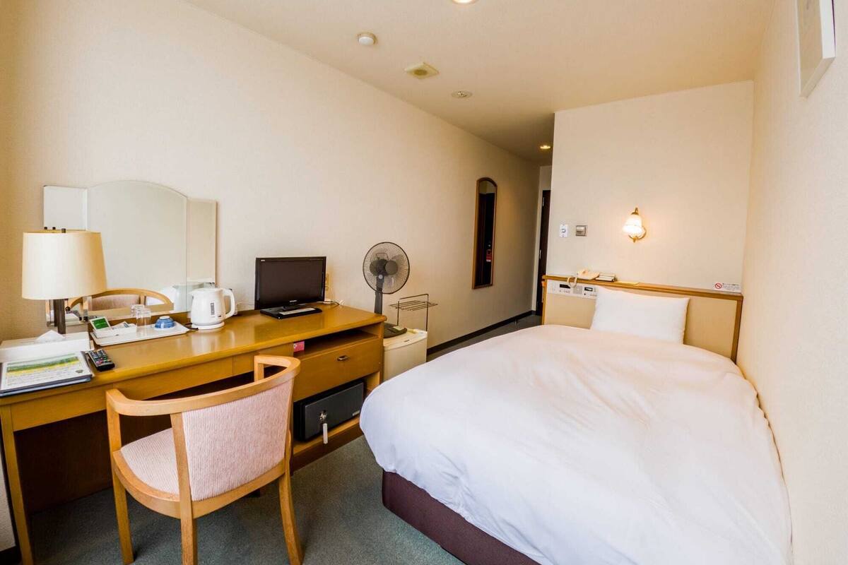 A hotel room with a bed, desk, chair, lamp, fan, and a bedside telephone.