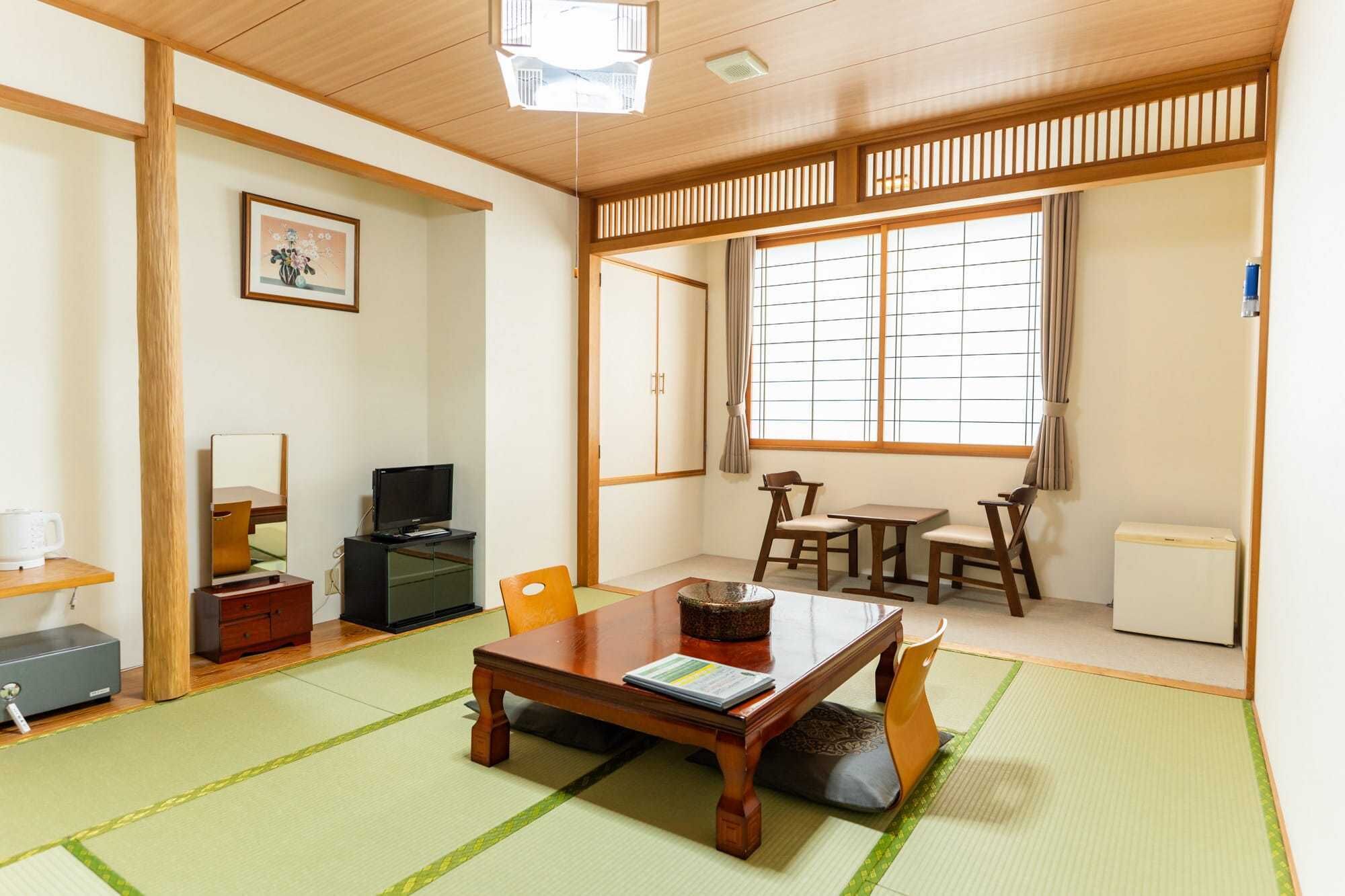 A traditional Japanese-style room with tatami flooring, a low table with cushions, a television, and a window with a view.