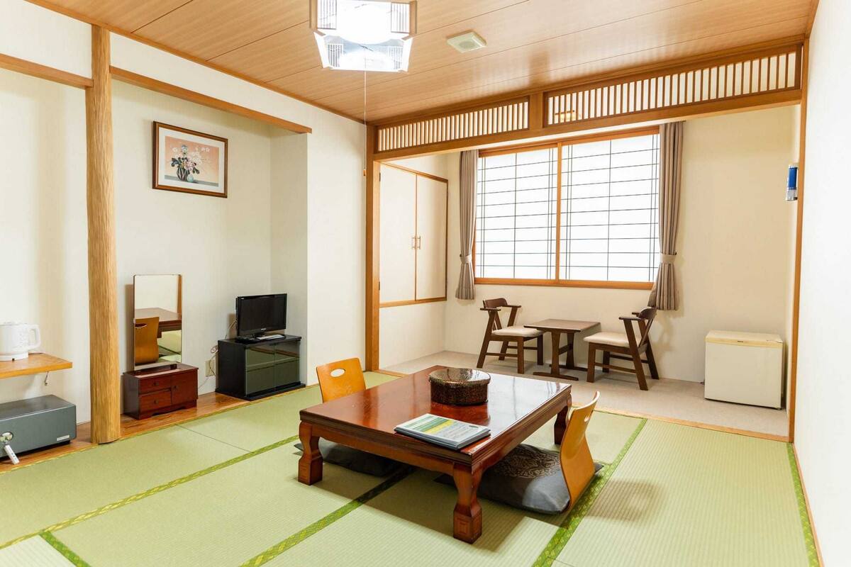 A traditional Japanese-style room with tatami flooring, a low table with cushions, a television, and a window with a view.